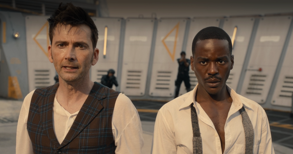 the Fourteenth and Fifteenth Doctor on top of a building, looking intently forward, with extras in the background