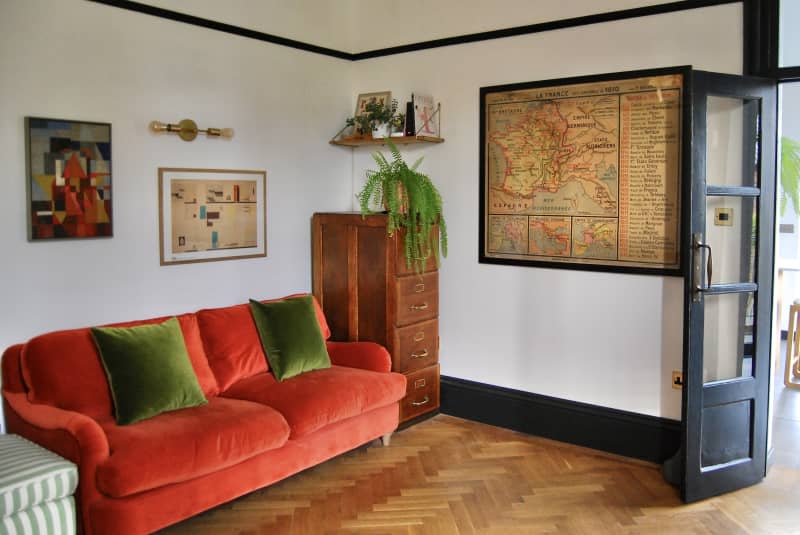 Red velvet love seat with green pillows next to wooden cabinets and walls with decorated artwork.