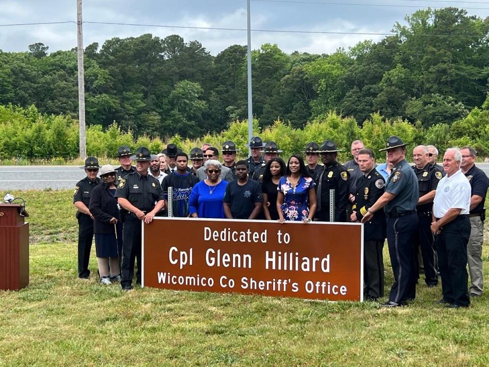 Members of the law enforcement community stand behind the Hilliard family and a sign dedicated to Cpl. Glenn Hilliard at a ceremony in Berlin, Maryland on June 12, 2023.