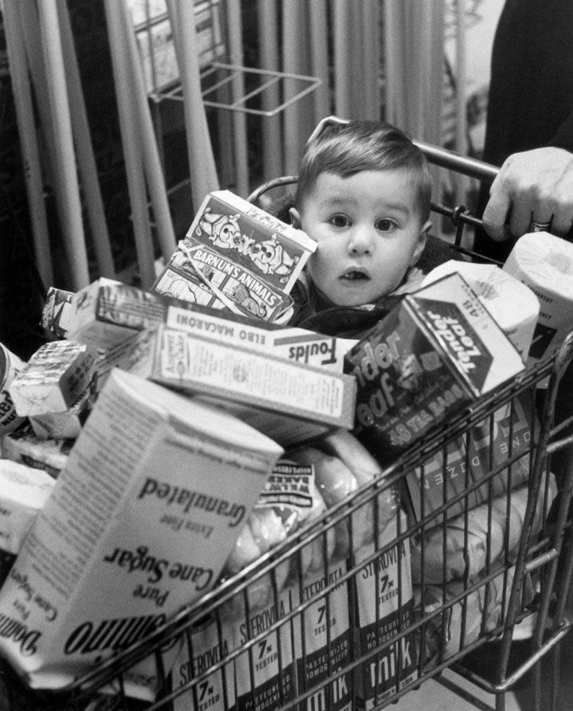 Niño dentro de un carrito de supermercado lleno de cajas con comida en la década de 1950.