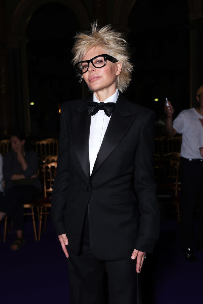 A person is wearing a sharp black tuxedo with a bow tie and large glasses, standing confidently and posing for the camera