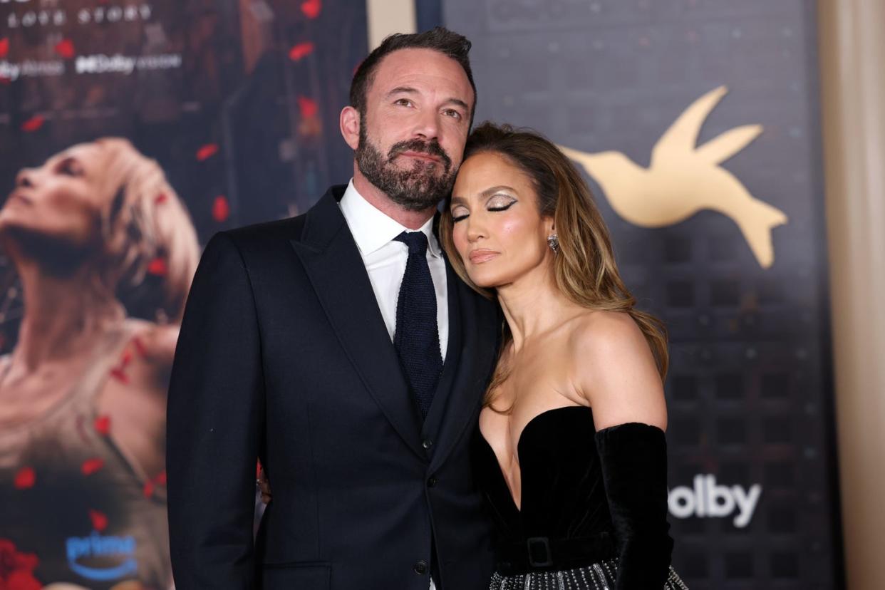 ben affleck and jennifer lopez hug while standing in front of posters, he wears a black suit jacket and tie with a white collared shirt, she wears a strapless plunging black dress with long black gloves, he looks up and smiles slightly, she closes her eyes and rests her head against him