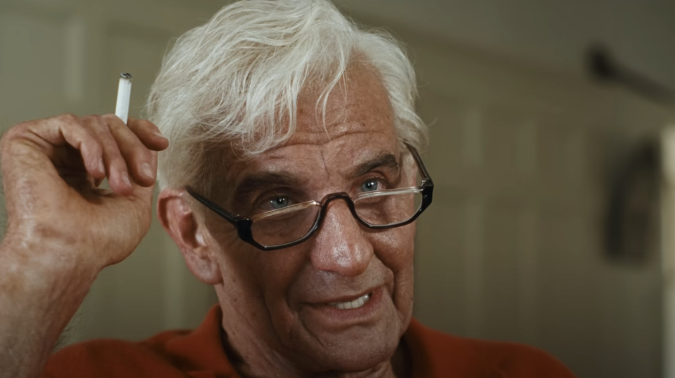 Leonard Bernstein with white hair, glasses, and a cigarette between his fingers, looking pensively off-camera in a scene from Maestro