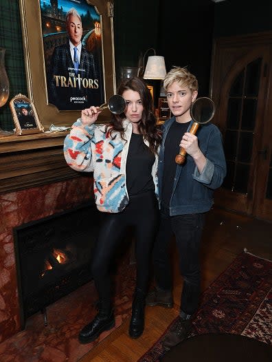 Megan Stalter and Sydney Lemmon pose in front of a framed "Traitors" poster, holding magnifying glasses, standing by a fireplace