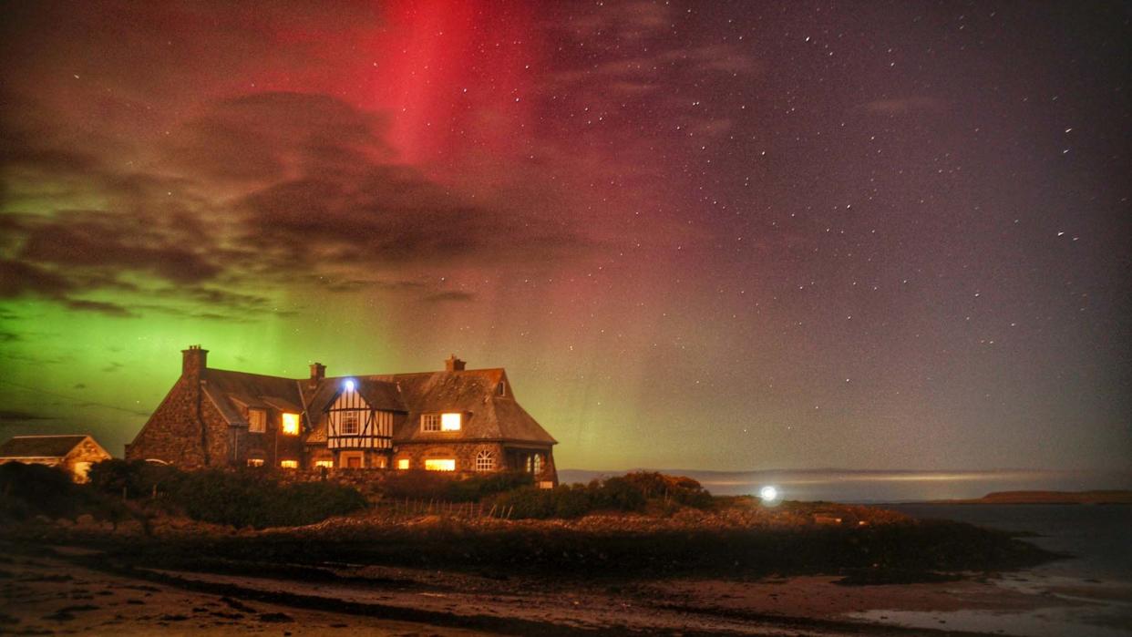  Green and red auroras shine over a lit-up house at night. 