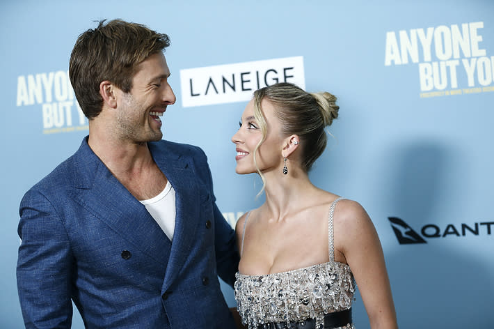 Two celebrities, a man and a woman, smile at each other. The man wears a blue suit; the woman wears a sparkling dress. They are at an event