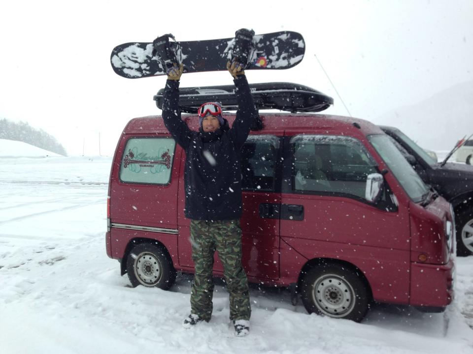 Man with snowboard in front of a tiny red van