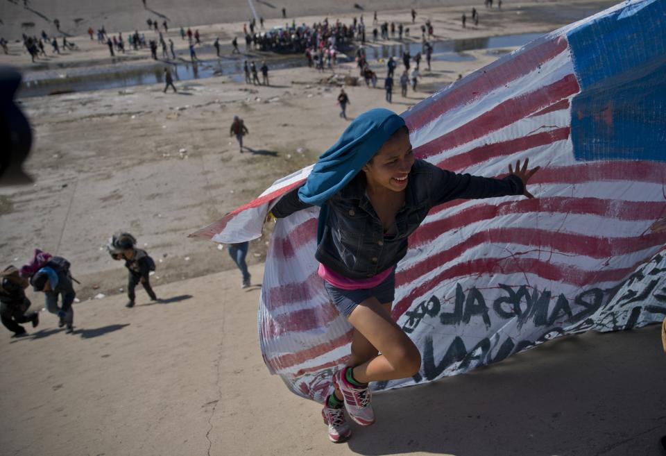 FILE - In this Nov. 25, 2018 file photo, a migrant woman helps carry a handmade U.S. flag up the riverbank at the Mexico-U.S. border after getting past Mexican police at the Chaparral border crossing in Tijuana, Mexico, as a group of migrants tries to reach the U.S. Activists, officials and social workers in Central America were staggered by the idea that U.S. President Donald Trump thinks he will help reduce immigration, by cutting off nearly $500 million in aid to Honduras, Guatemala and El Salvador; exactly the opposite will happen, they say. (AP Photo/Ramon Espinosa, File)