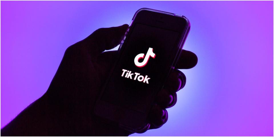 Hand holding a phone with a TikTok logo on the screen. Background is purple.