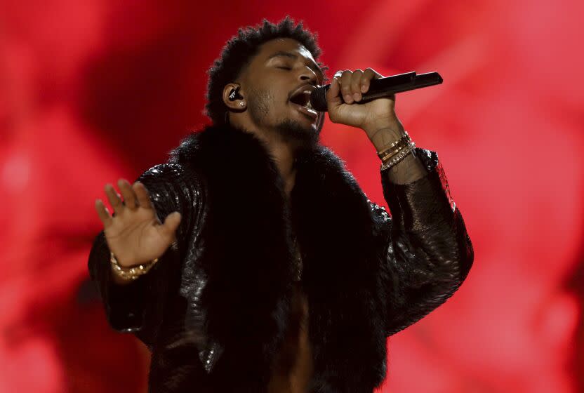 A man with short black hair wearing a black outfit and singing into a microphone against a red background