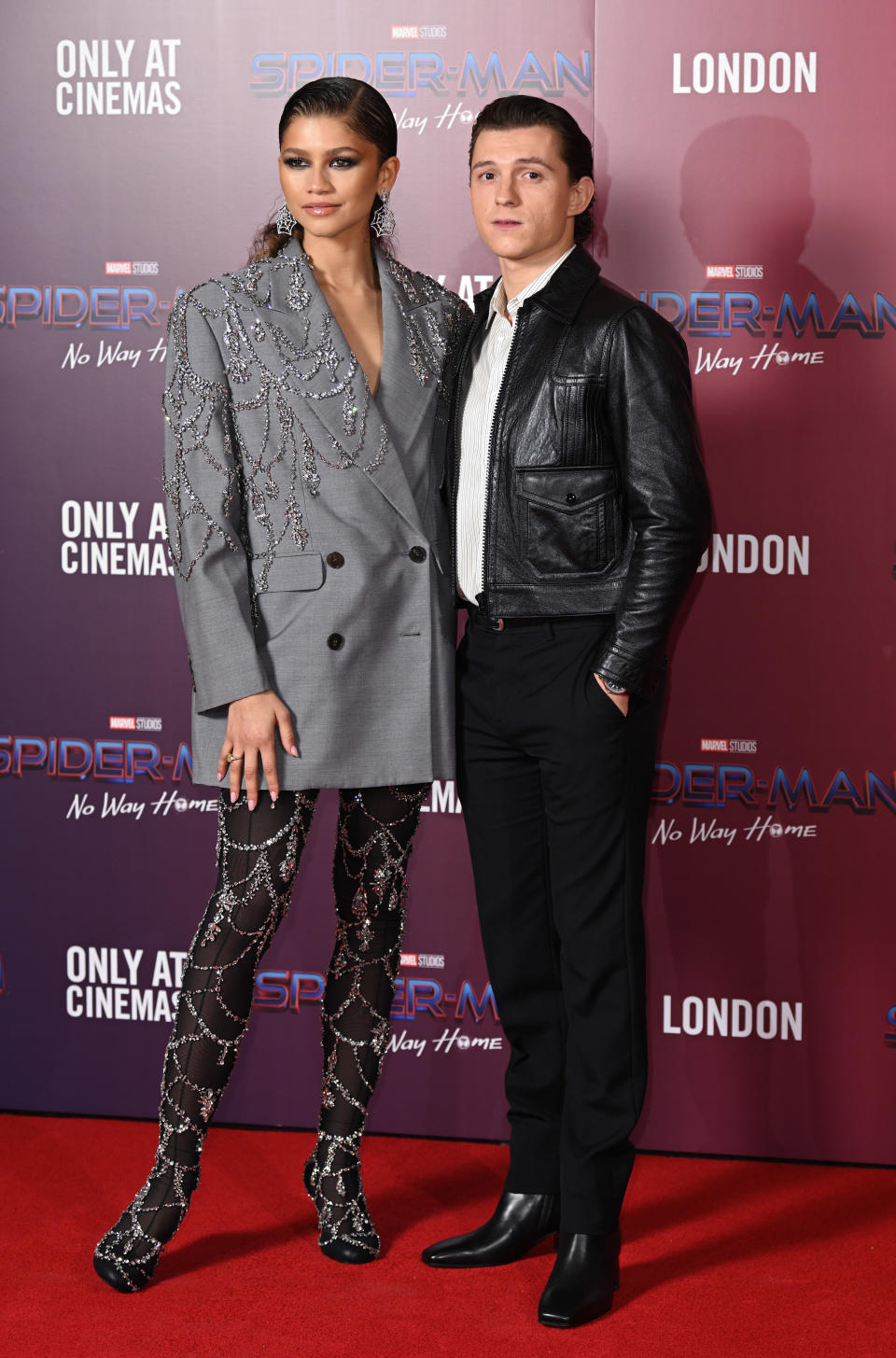 Zendaya wearing an oversized jacket as a shirt with jewel details and matching leggings/boots next to Tom who is wearing a leather jacket and slacks