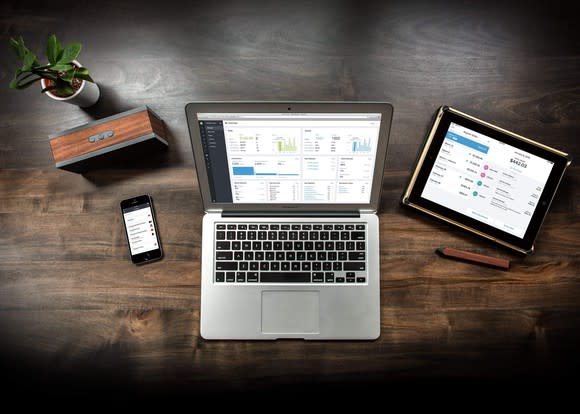 A laptop, a cellphone, and a tablet on a wooden desk, all showing the Shopify app.