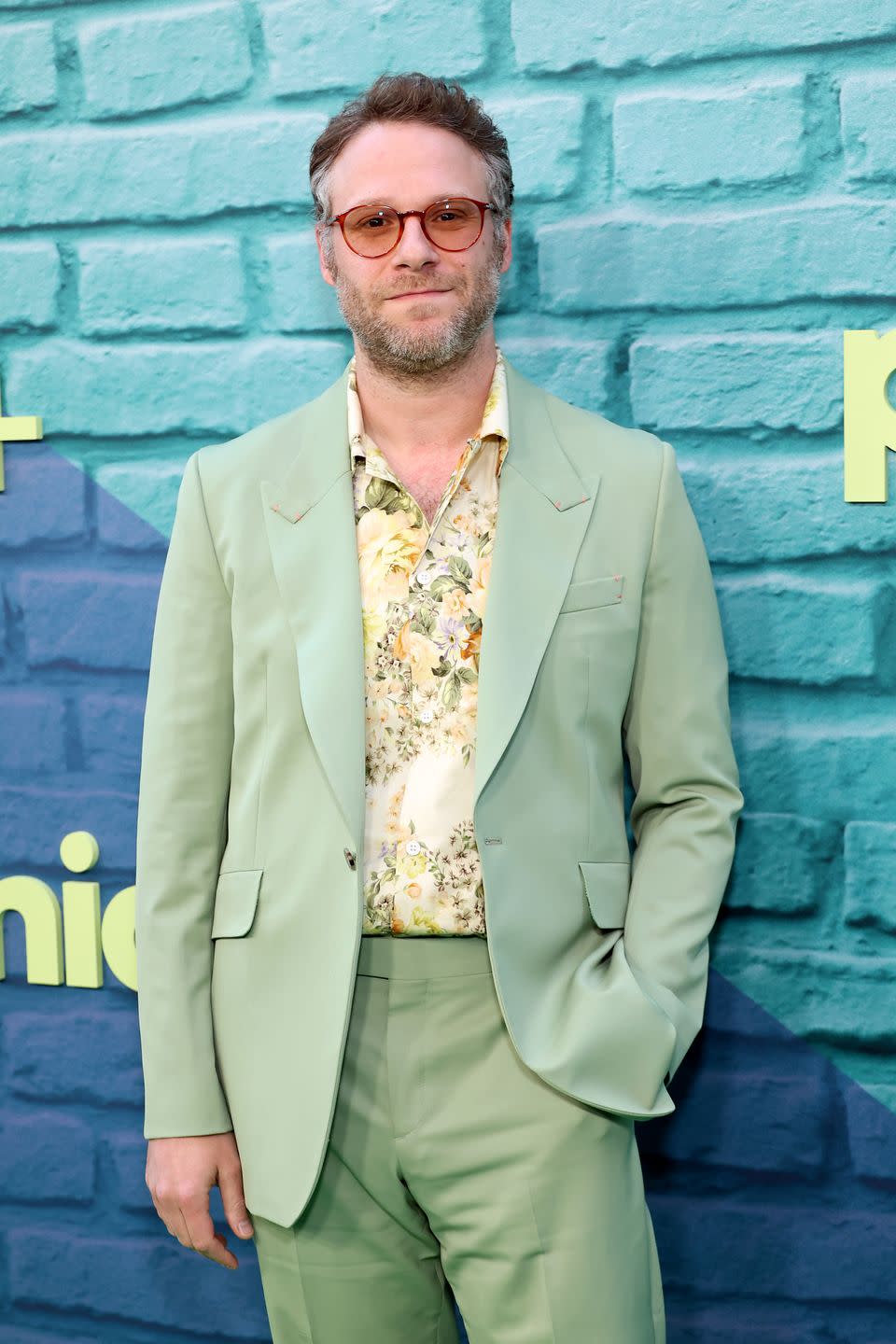 seth rogen in a light green suit and orange tinted glasses smiling against a turquoise brick background