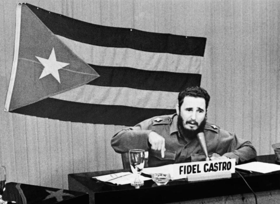 Fidel Castro speaks at a press conference, gesturing with his hand while sitting at a table. The Cuban flag is displayed in the background