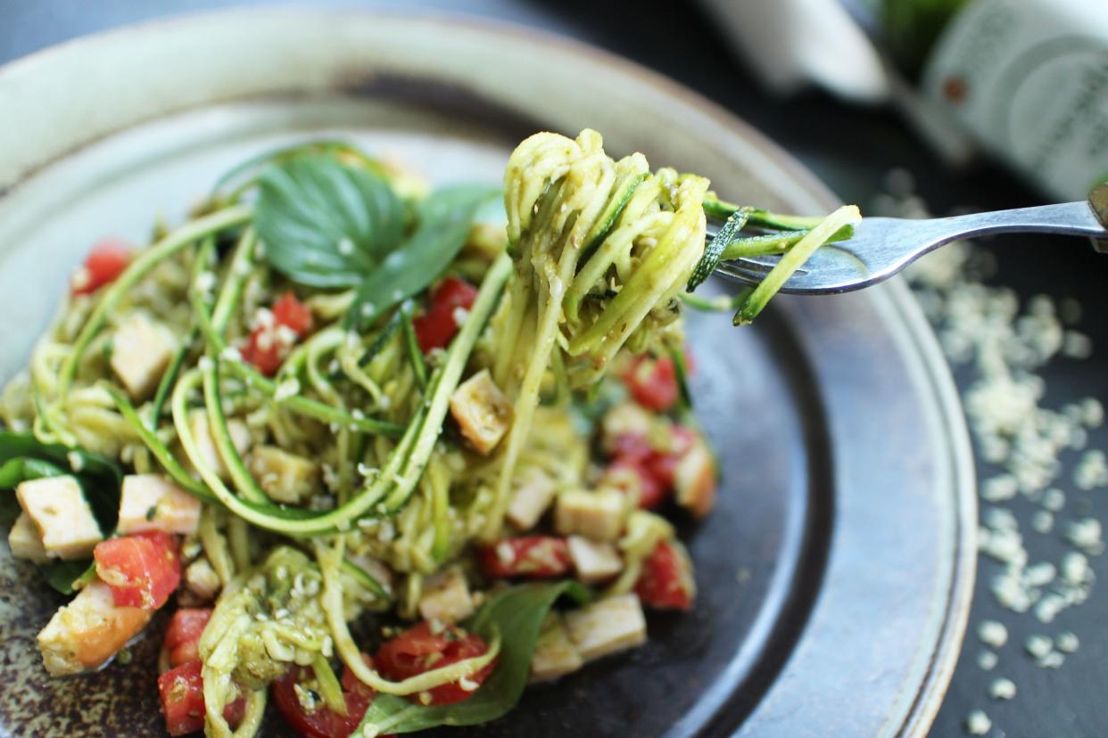 Closeup of Heather's zucchini noodles with basil-pumpkin seed pesto on a fork over an artisanal stainless steel plate, selective focus, on a dark brown table with white seeds, blurred