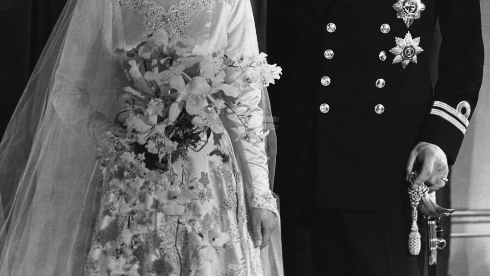 princess elizabeth and philip mountbatten stand and look at each other smiling, she wears a wedding dress, veil and crown and holds a bouquet, he wears a dark military uniform and holds a sword