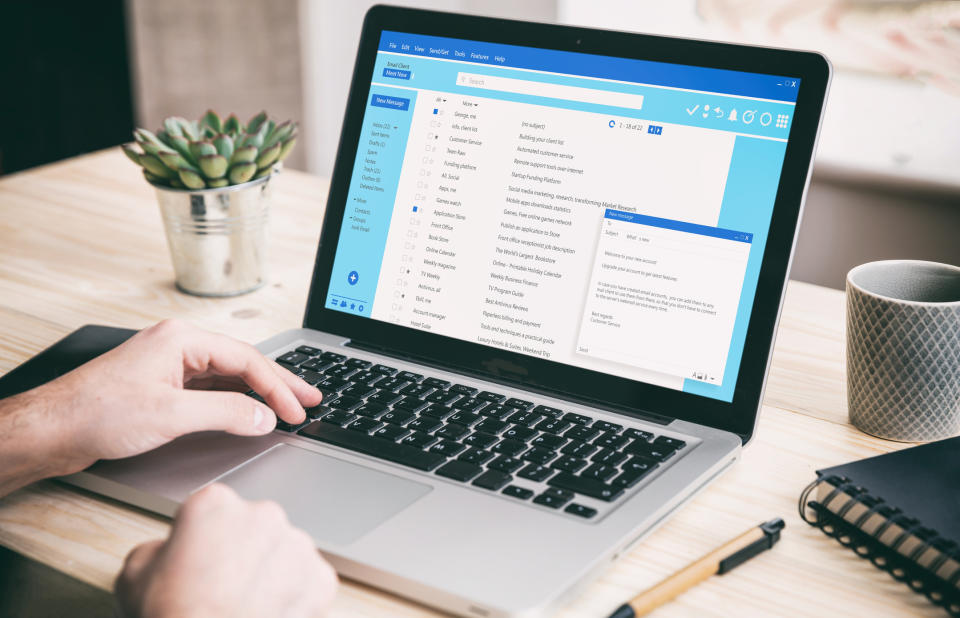 Person typing on a laptop, with an email application open on the screen. A small plant and a spiral notebook are on the desk