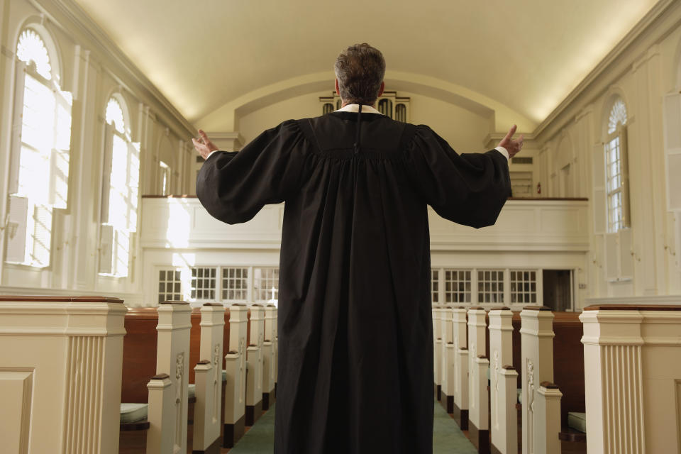 A man in a black robe stands at the front of a church with arms raised, facing empty pews and a pipe organ in the background