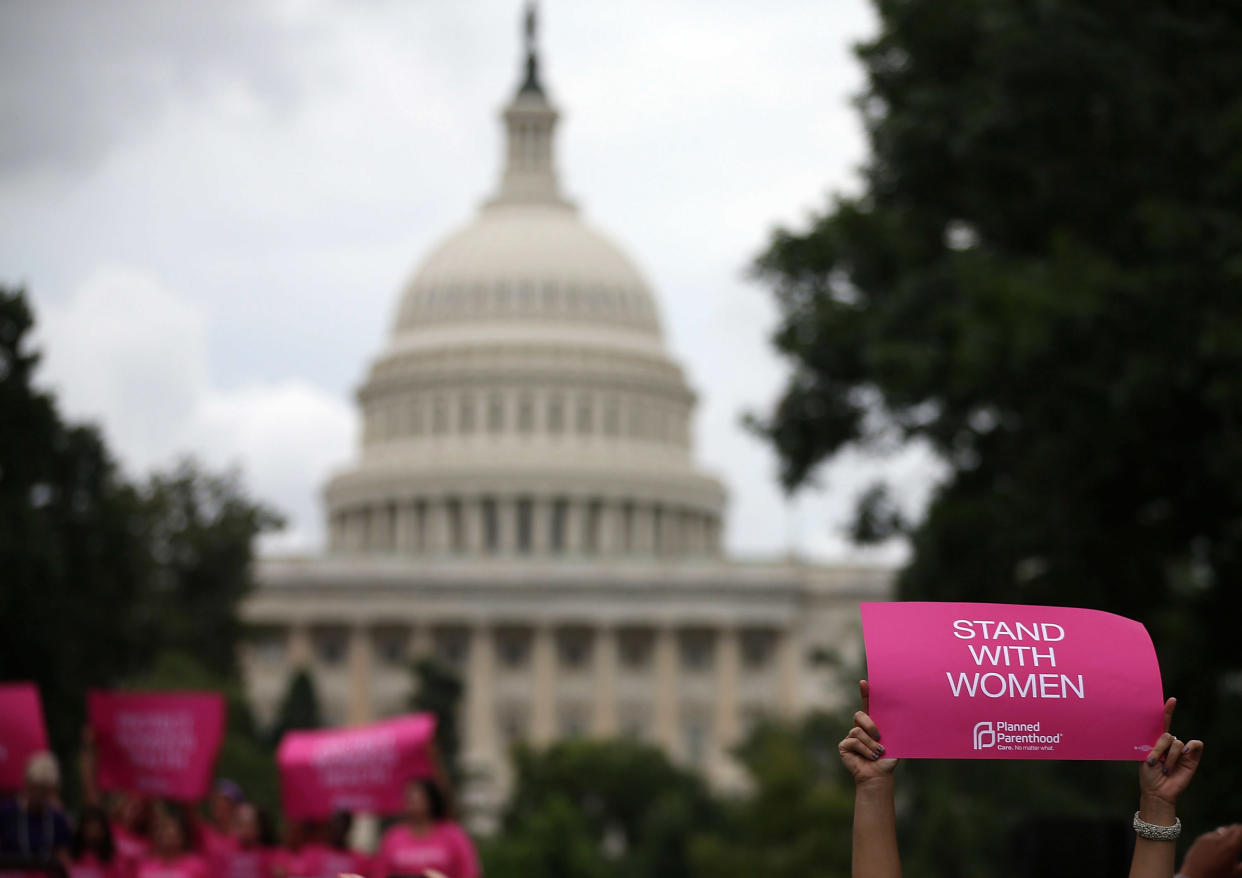 Women in front of the Capitol hold up pink signs saying: Stand With Women.