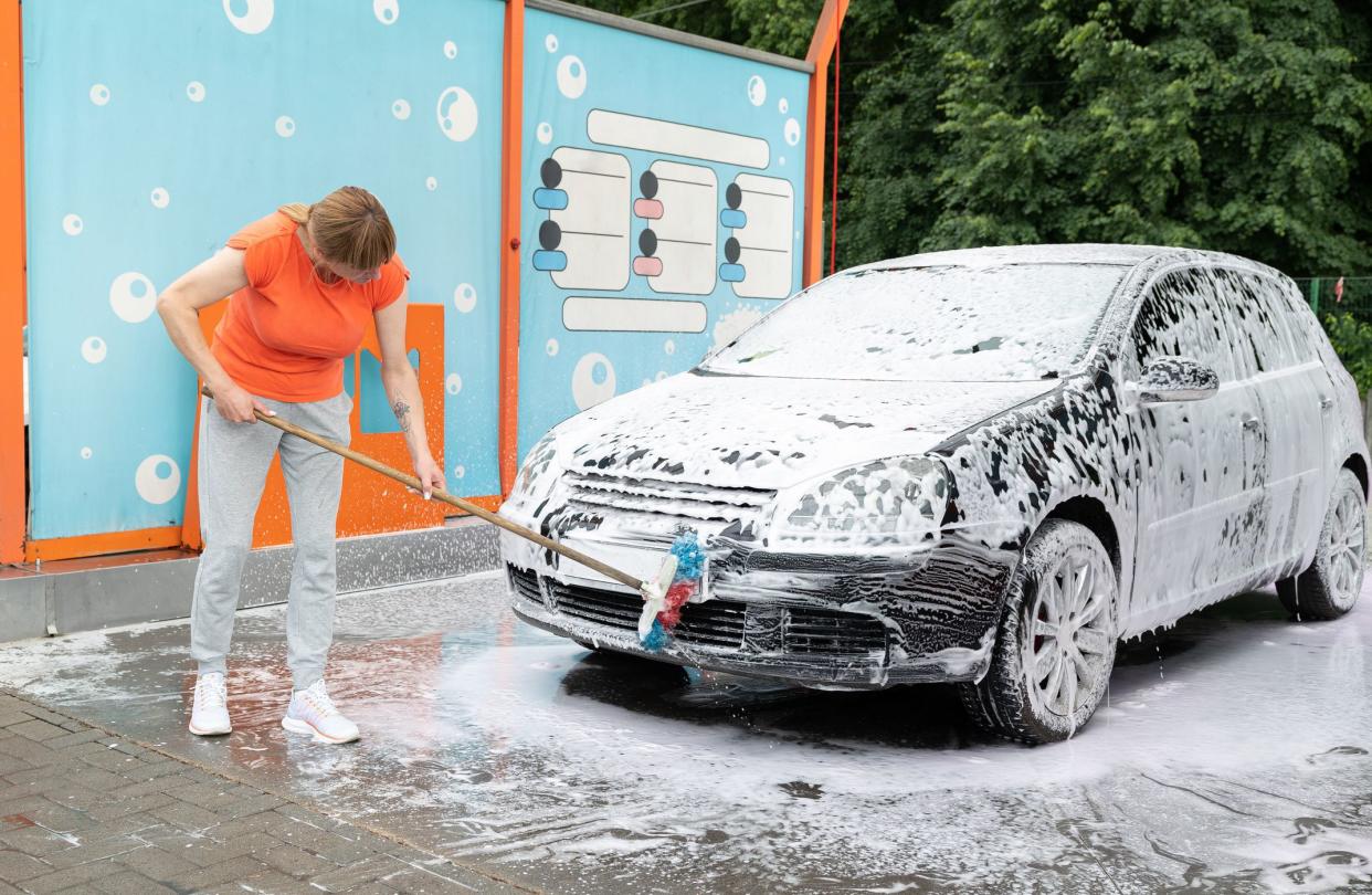 The woman washes her car at a self-service car wash. The car owner uses a long-handled brush to wash his car. The car is covered with washing foam.