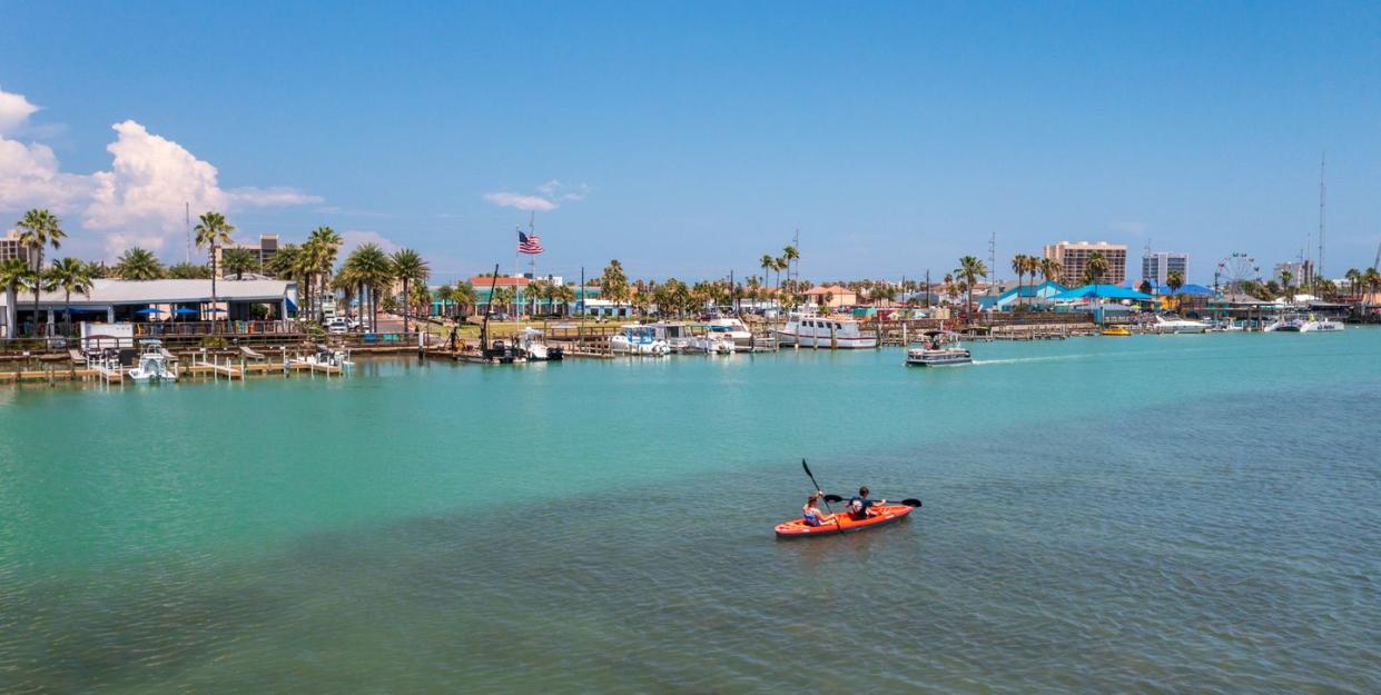 person kayaking on the ocean next to the shore covered with palm trees, boats, businesses and more