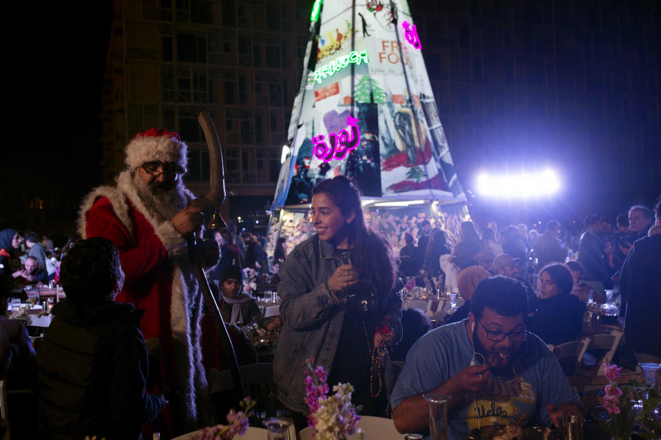 In this Monday, Dec. 23, 2019 photo, a volunteer, center, serves drinks at a public Christmas dinner as a boy asks Santa Claus for a selfie, in Martyrs Square where anti-government activists are encamped, in Beirut, Lebanon. As Lebanon's protest movement enters its third month, the economic pinch is hurting everyone. But Lebanese are resorting to what they've done in previous wars and crises: They rely on each other, not the state. (AP Photo/Maya Alleruzzo)