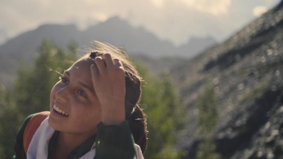 A young girl smiles while adjusting her hair against a mountainous backdrop; still from 'Yasmeen's Element'