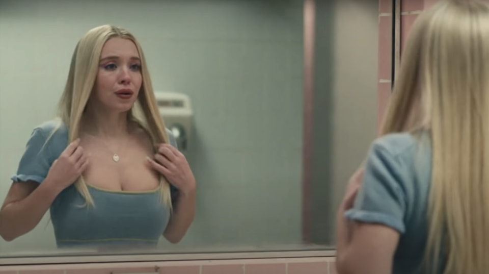 Sydney Sweeney, standing in front of a mirror, appears emotional, adjusting her dress with her hands. She looks at her reflection in a bathroom