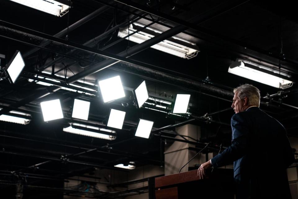 A man in a suit speaks at a lectern while bright lights shine down on him.