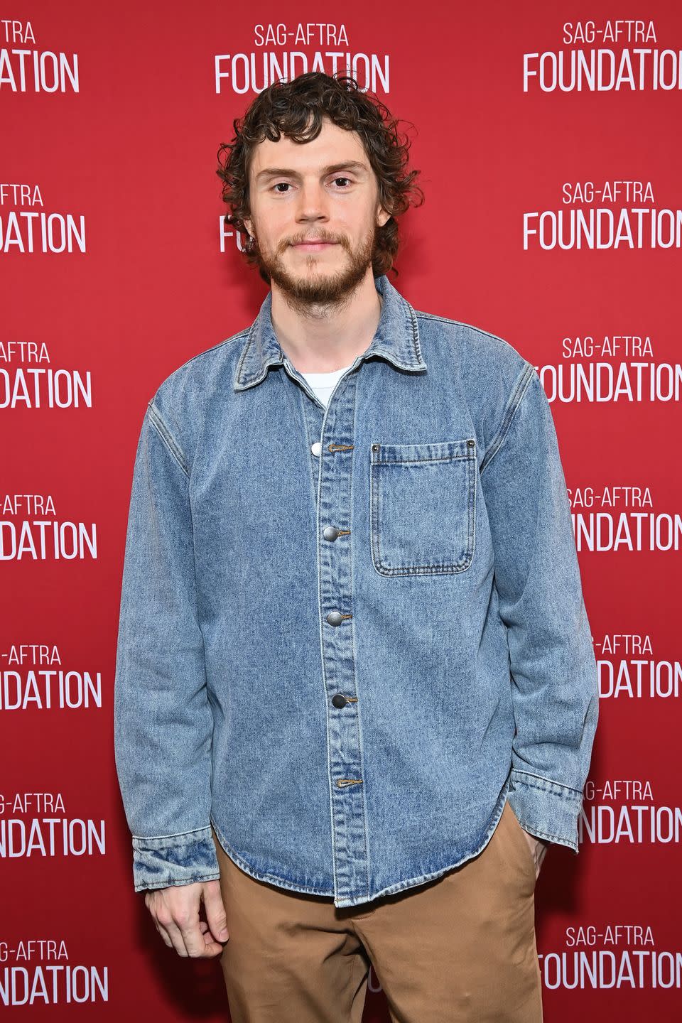 evan peters with a beard wearing a denim shirt and smiling in front of a red background