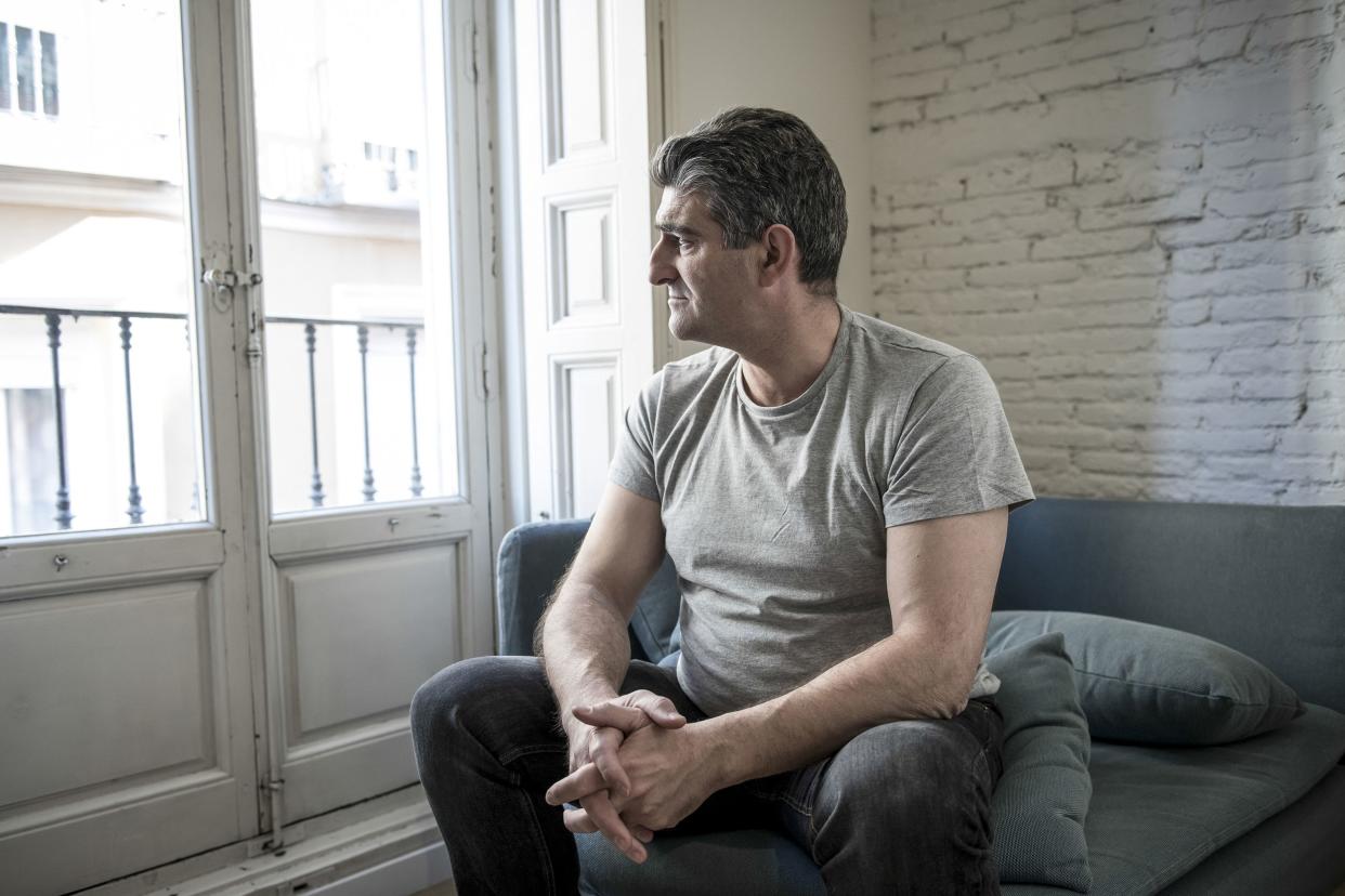 Worried middle-aged man sitting on a dark grey couch in an apartment, his head is looking out of the balcony windows on the left, a white painted brick wall on the right background