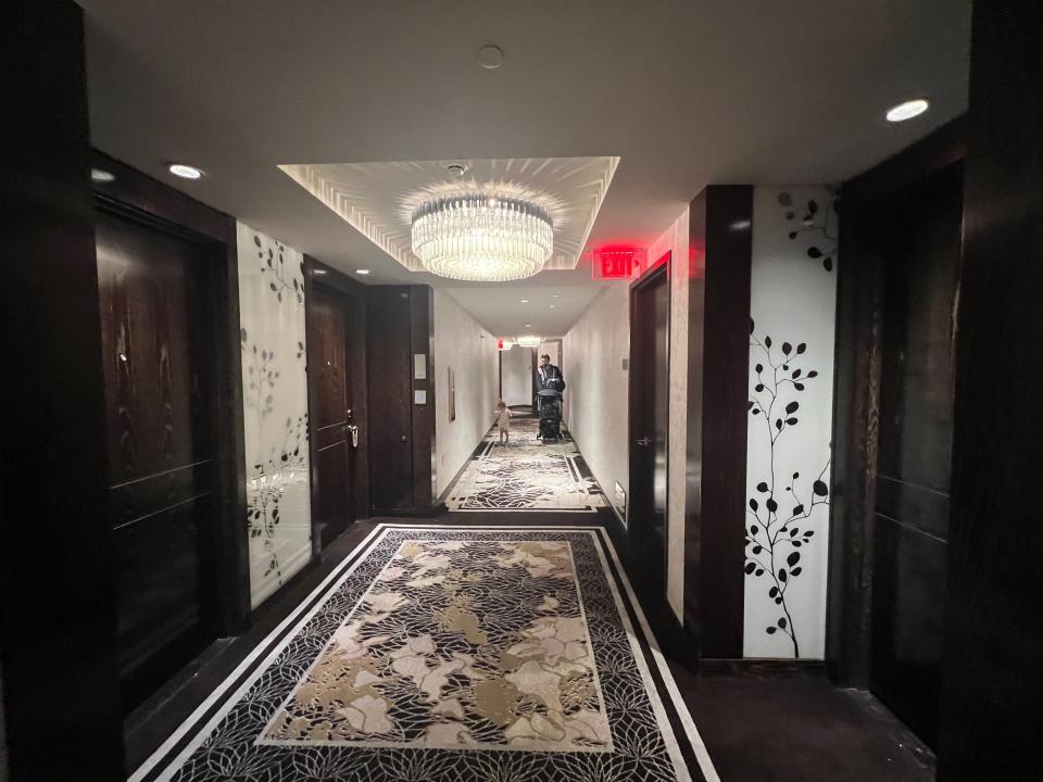 A long hotel hallway with a chandelier and a man and his child walking down it.