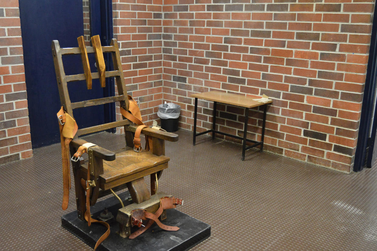 The 110-year-old electric chair, with its wooden head rest and orange restraints, in a plain brick room that also contains a small table and a trash can lined with plastic. 