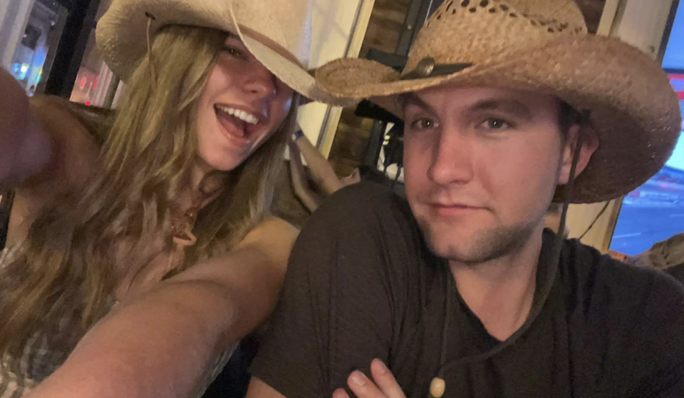 Two people wearing cowboy hats smiling for a selfie, indoors with faint background view of a sports arena