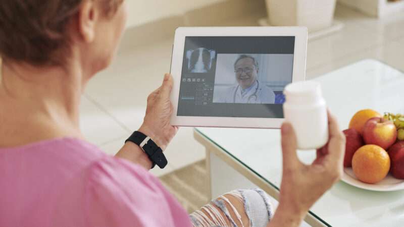 A woman holds a bottle of pills while speaking to a doctor on a tablet over telemedicine.