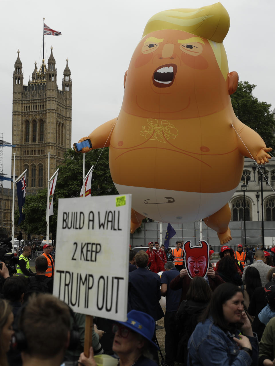 FILE - In this June 4, 2019 file photo, a woman holds up a banner standing next to the 'Trump Baby' blimp as people gather to demonstrate against the state visit of President Donald Trump in Parliament Square, central London. President Donald Trump is being trolled by an angry diaper-clad caricature armed with a cell phone. It’s Baby Trump, the blimp that has become synonymous with resistance to the American president. The balloon has been cloned multiples times over and become something of a celebrity _ for at least one slice of the U.S. electorate. He’s also emerged as a rallying point for supporters of the president who see the blimp as evidence of just how over-the-top the opposition has become. (AP Photo/Matt Dunham)