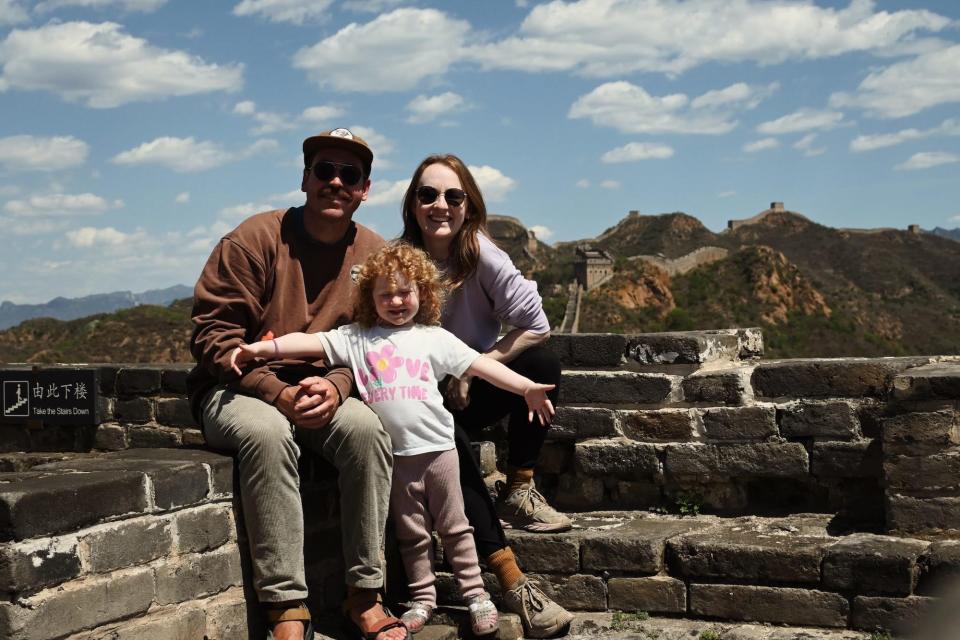 A couple and their daughter pose on a brick wall with the Great Wall of China behind them.