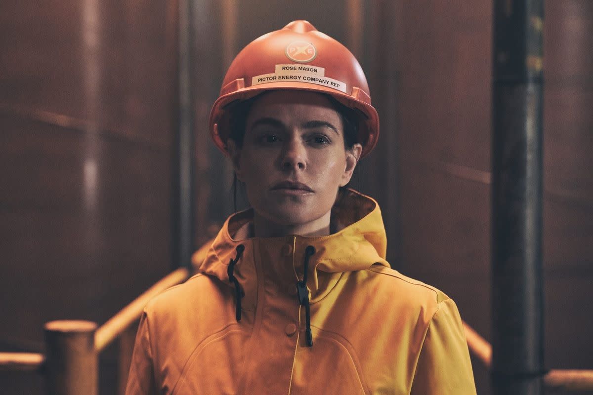 emily hampshire in the rig, wearing a hard hat and hi vis and looking glum