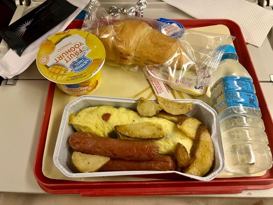 Breakfast meal with eggs, potatoes, sausage, yogurt, and croissant on tray.