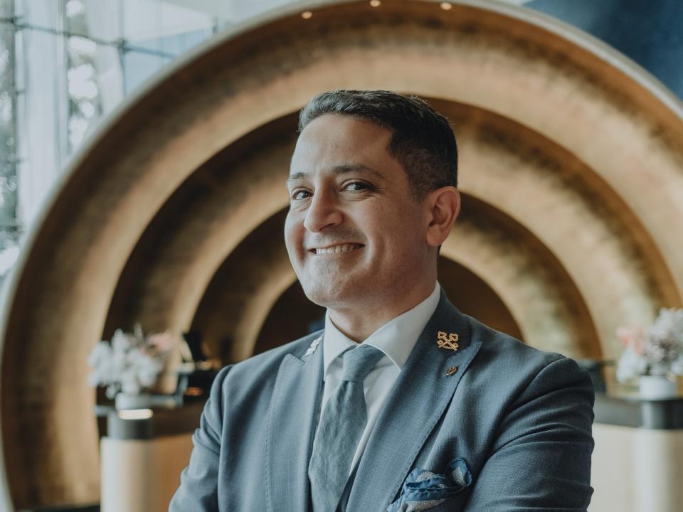 A man in a gray suit stands smiling in a luxurious hotel lobby