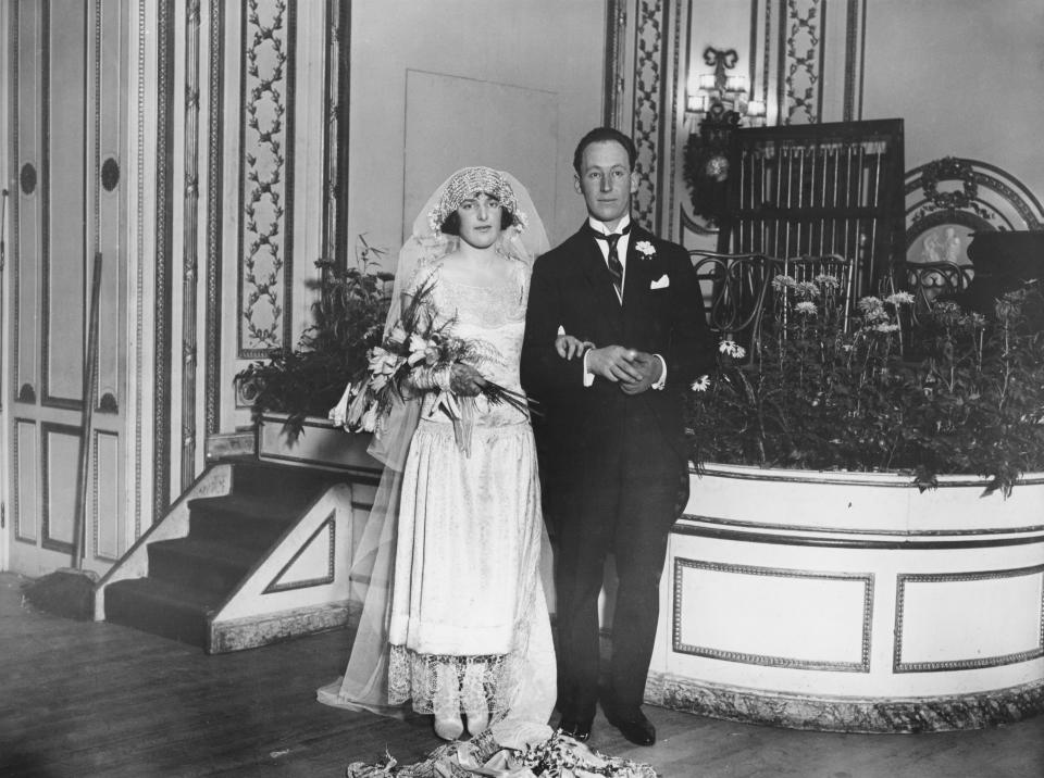 Bride and groom in vintage wedding attire, 1920s setting, standing arm-in-arm in an ornate room with floral arrangements in the background. Names unknown