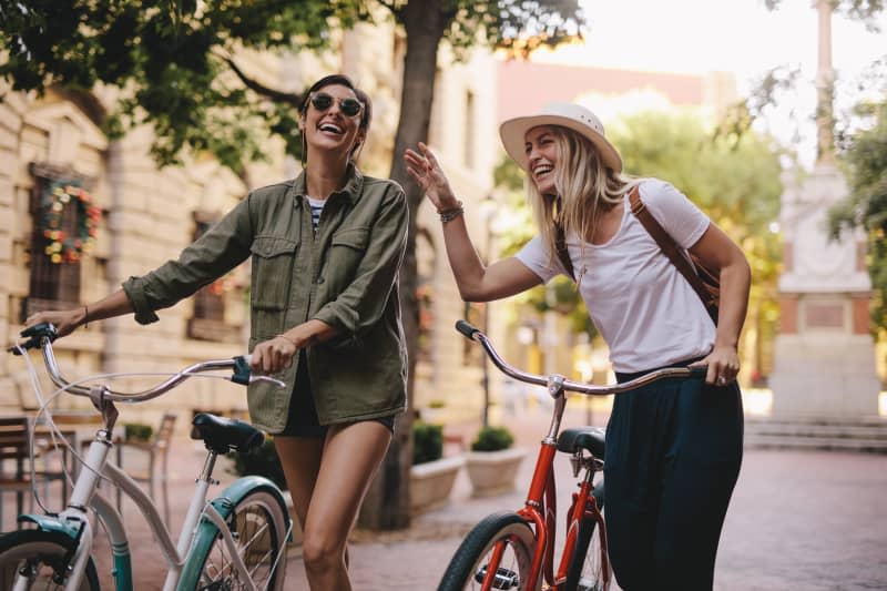 Positive and happy girls walking on the city street with bicycles. Female friends enjoying a walking down the street with their bikes.