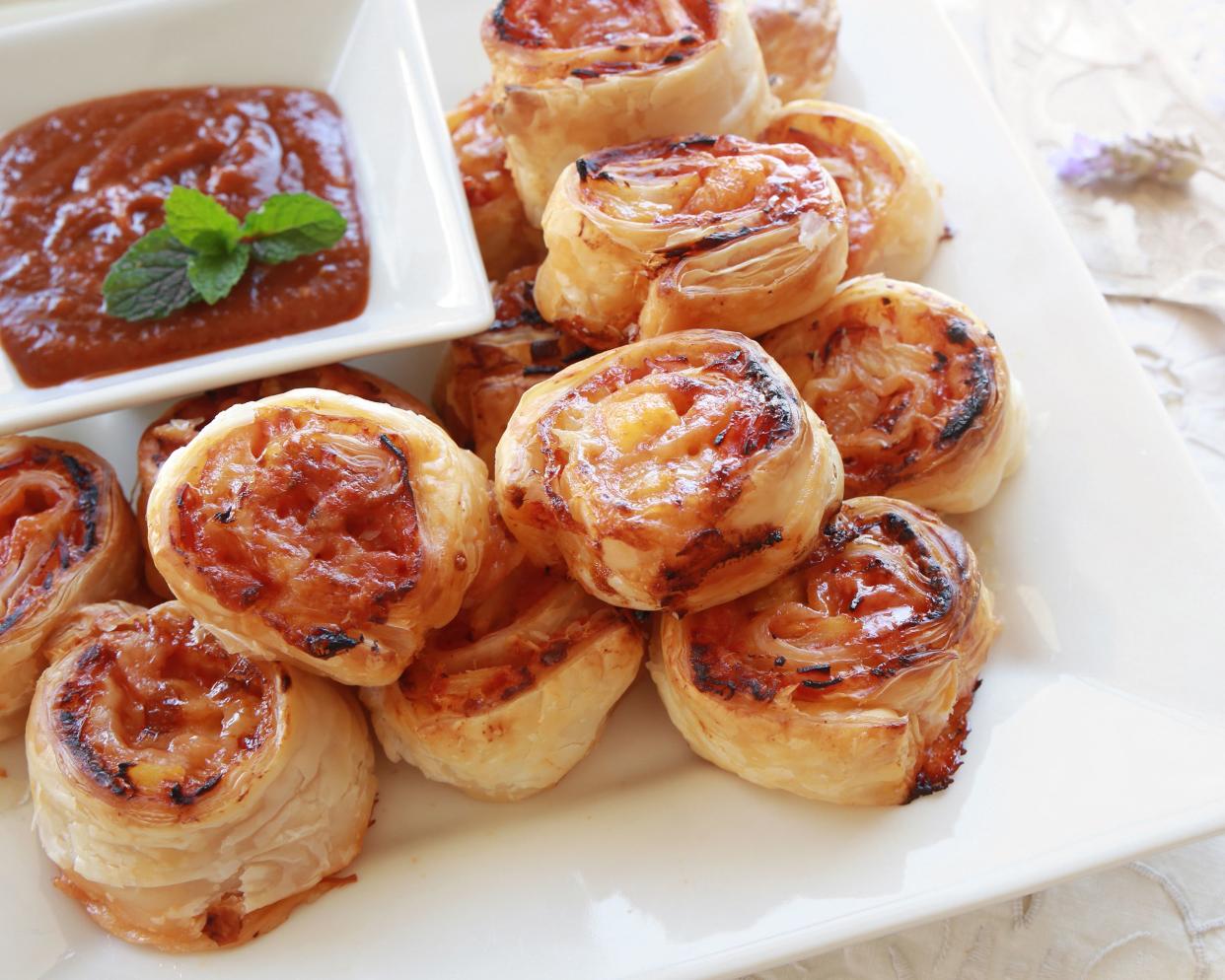 Ham and cheese wheels on a white square plate with a tiny white square plate with marinara sauce on a natural colored table