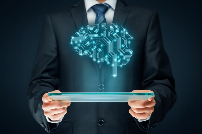A man in a suit holding a tablet. An illustrated brain made of electrical connections hovers above the scree, illustrating artificial intelligence.