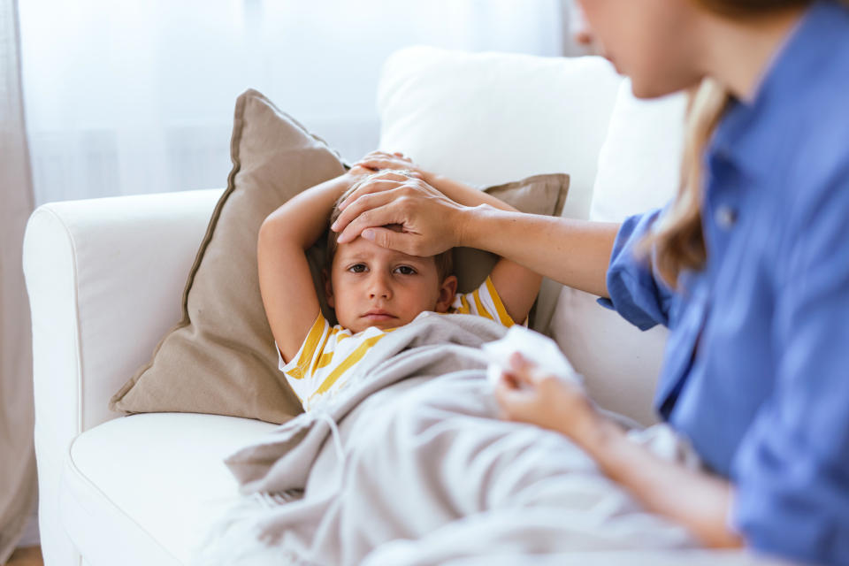 A loving mother is seen tending to her sick child on a white sofa, gently checking his temperature and ensuring his comfort in a cozy, well-lit living room environment.