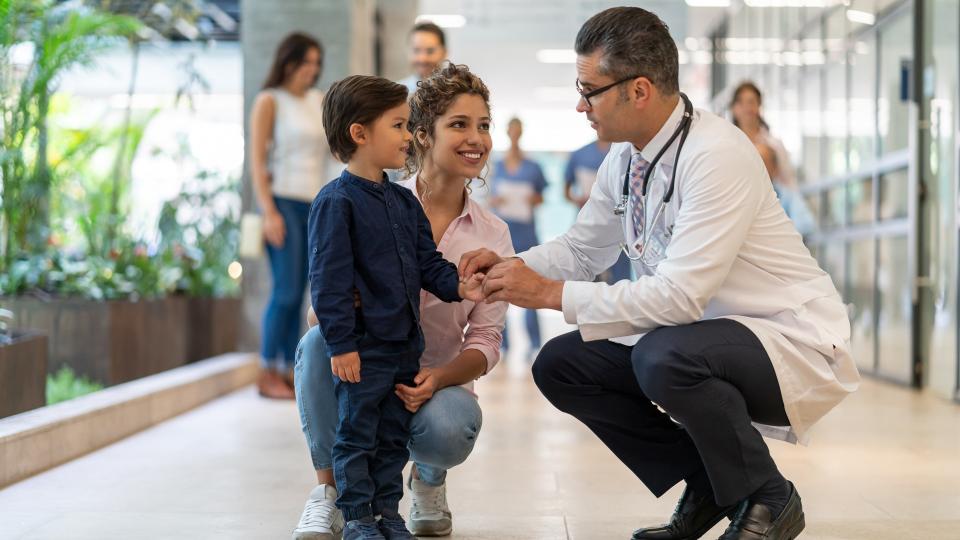 Male pediatrician talking to his little patient who is standing next to his mom all smiling - Incidental people at background.