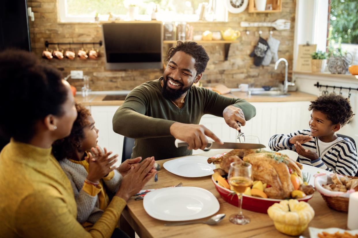 Happy African American man having Thanksgiving lunch with his family and carving stuffed turkey at dining table.