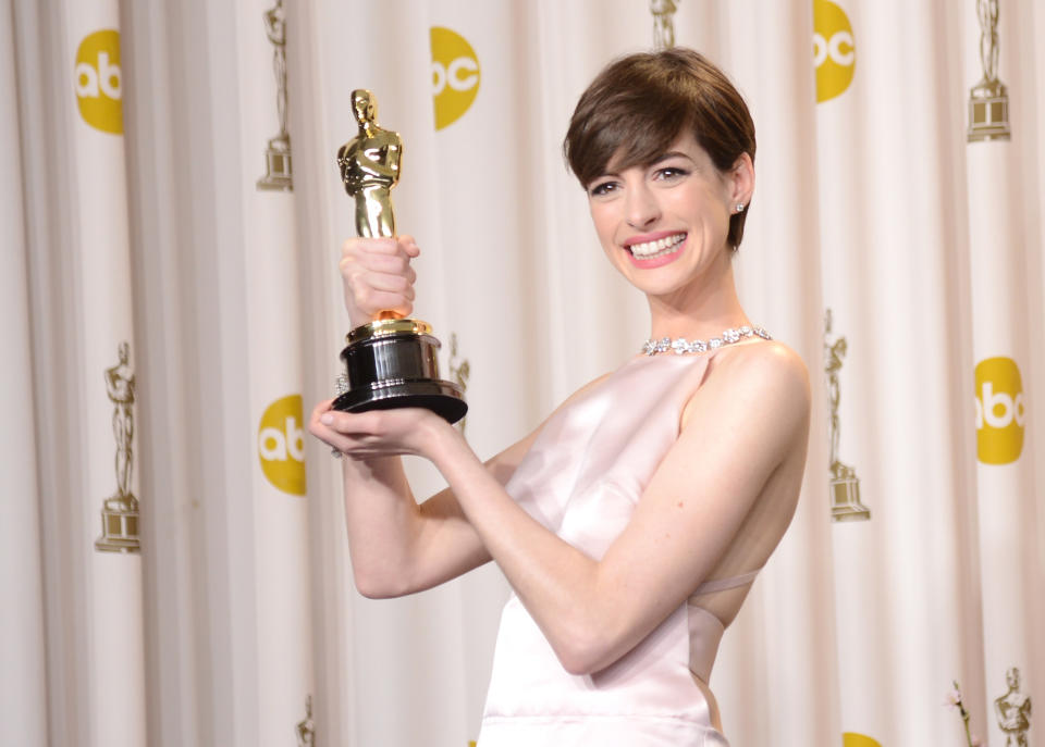 Anne Hathaway in a sleek gown holding an Oscar trophy, smiling at the camera