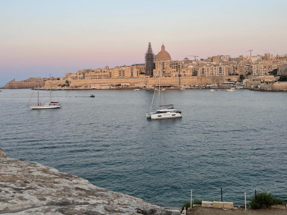 Photo of a harbor with several ships moored in the water. The photo is taken from a stone wall, which the viewer can see at the bottom of the photo. On the horizon is the Malta cityscape.