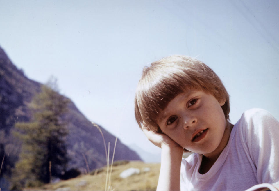 A young child with short hair and a white shirt leans their head on their hand, posing outdoors with mountains in the background